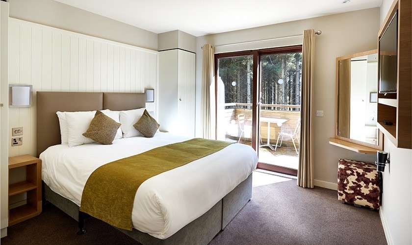 Large double bed sits neatly made with white linens and green runner; sliding glass doors open to a balcony with chairs overlooking pine trees, within a tidy, modern bedroom.