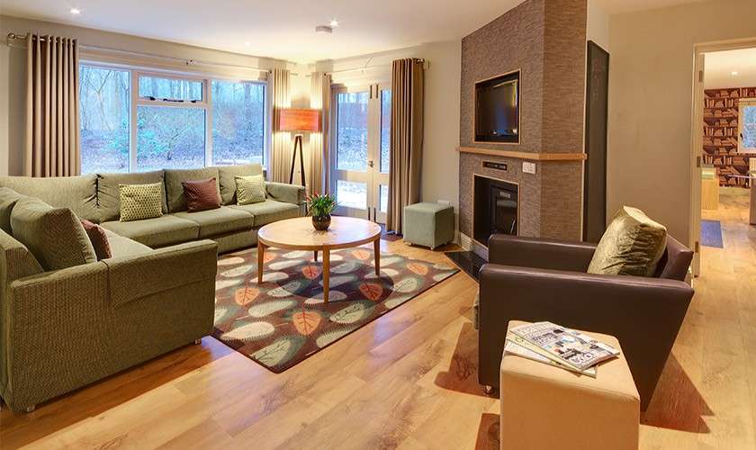 Sectional sofa surrounds a round coffee table, facing a TV above a fireplace; floor lamp glows, armchair and ottoman sit on leaf-patterned rug in a wood-floored, windowed living room.