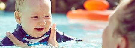 Baby smiles while being gently held under the arms, bobbing in shallow pool water; nearby adult faces them, sunlit outdoor setting with soft focus background and an orange float.