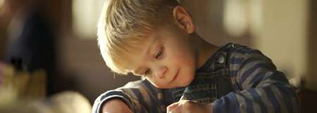 Child concentrates, drawing with a pencil on paper, sitting at a table indoors. Warm light from a window illuminates their face and striped shirt, with background softly blurred.