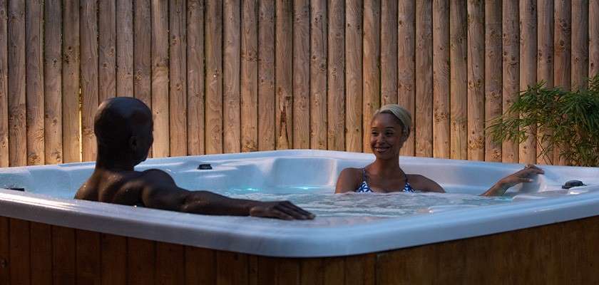 Two adults relax and chat in a bubbling outdoor hot tub, leaning on the rim. Wooden privacy fence surrounds them, evening lighting glows softly, with a potted plant nearby.