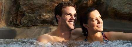 Couple relaxing—two adults lean back in a bubbling hot tub, smiling and gazing upward; context: natural stone backdrop with warm light suggesting an outdoor spa setting.