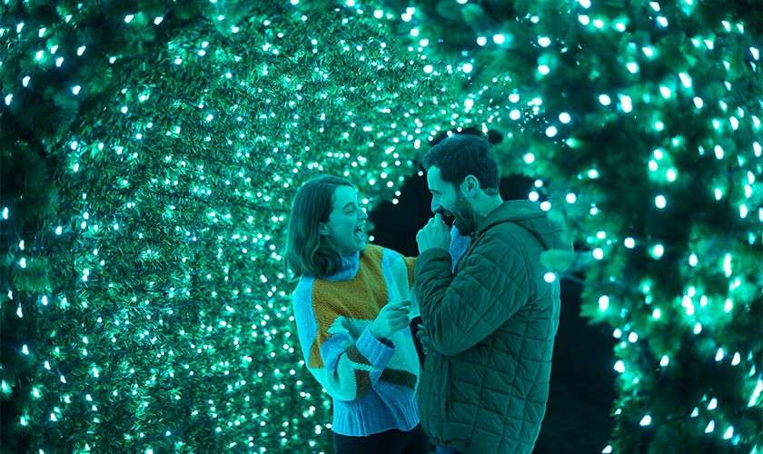 Two people laugh and chat, facing each other, inside a glowing tunnel of green-blue string lights. The illuminated arch surrounds them densely, suggesting a festive nighttime light display.