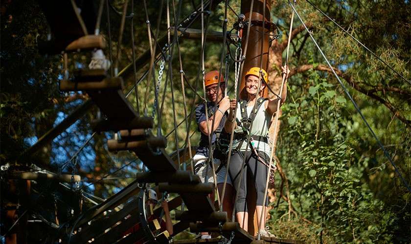 Two helmeted climbers hold safety lines while crossing a suspended rope bridge, navigating platforms and cables within a forested treetop adventure course.