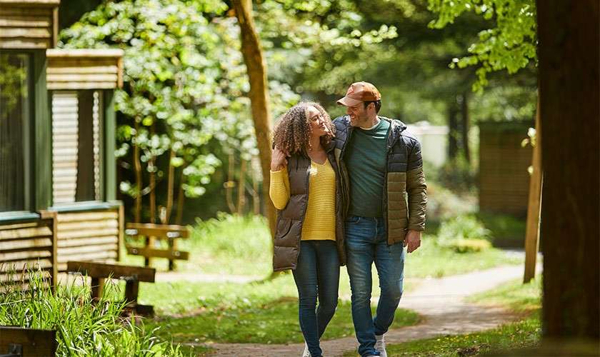 Couple walk arm-in-arm, smiling and looking at each other, along a sunlit, tree-lined path. Wooden cabins and benches line the green, leafy park setting with soft shadows and winding walkway.