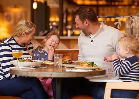 Two adults and two children share a meal, laughing together. They sit around a round table with salad, burgers, fries, and drinks, in a warm, softly lit restaurant.