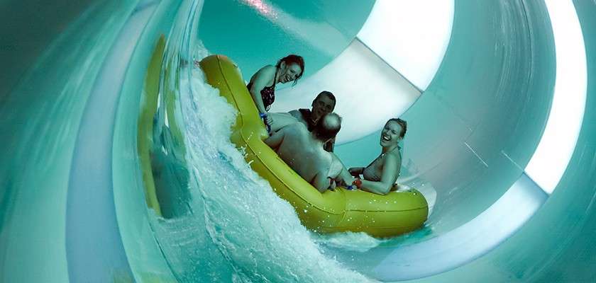 Four swimsuit-clad riders steer a yellow inflatable raft, rushing through an enclosed, turquoise-lit water slide, with splashes and curved tube walls surrounding them.