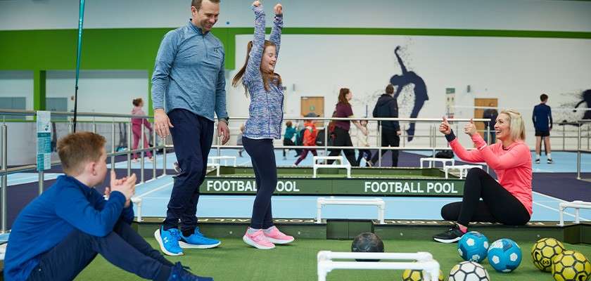 Girl raises arms celebrating while adults and a boy watch; soccer balls sit on a small football pool table in an indoor recreation center. Text: FOOTBALL POOL, FOOTBALL POOL.
