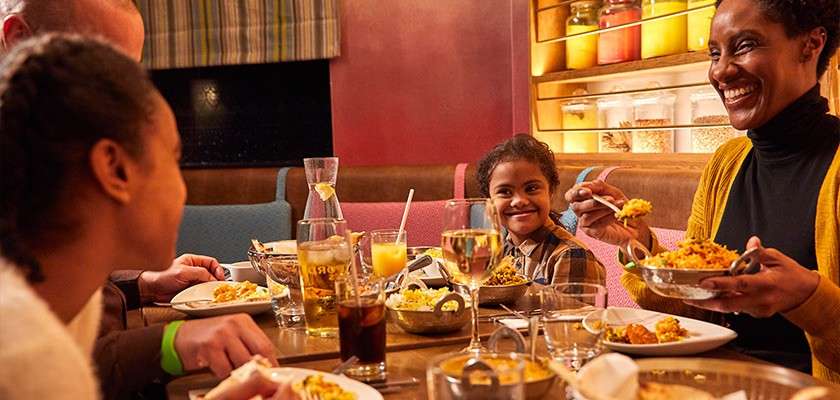 Family diners eat spoonfuls of rice and curry, smiling and talking. Plates, glasses, and bowls fill the table. Warm restaurant setting with booths and shelves of jars lit behind.