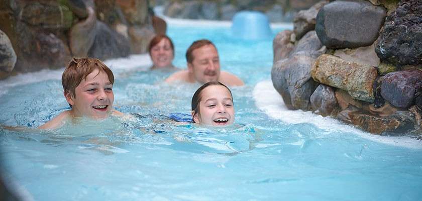 Two children swim ahead, smiling, followed by two adults, as they float through a lazy river. Water churns gently against rock-lined walls in a bright, indoor aquatic setting.