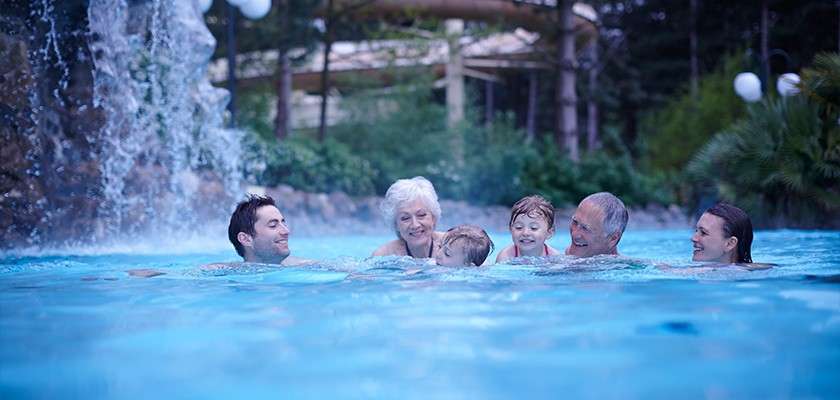Group of six people swim and smile together, facing each other. Bright blue outdoor pool with a nearby rock waterfall, lush trees, and wooden structures in the background.