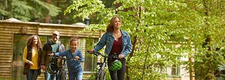 Two cyclists walk their bikes along a leafy forest path; a child pushes another bike beside them while two adults follow; wooden cabins and tall trees surround the trail.