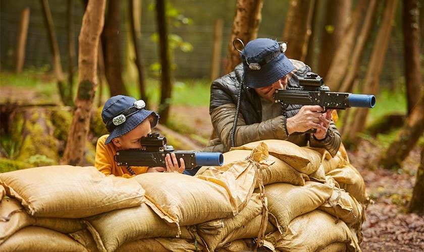 Two players aim laser tag rifles while crouching behind stacked sandbags; both wear hats with sensors. They take cover in a wooded outdoor arena with trees and fencing in background.