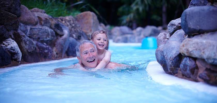 An older man carries a child on his back, smiling, as they float through a lazy-river pool surrounded by rock walls and tropical plants.