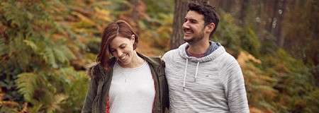 Two adults walk side by side, smiling, in casual jacket and hoodie, through a forest with ferns and tall trees, warm autumn colors surrounding them.