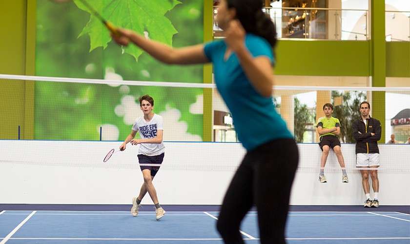 Badminton players rally; a person in teal swings, while a boy prepares to return across the net; two spectators watch courtside in a bright indoor gym with a green leaf mural. Shirt text: “RIDE [illegible] WAVE”.