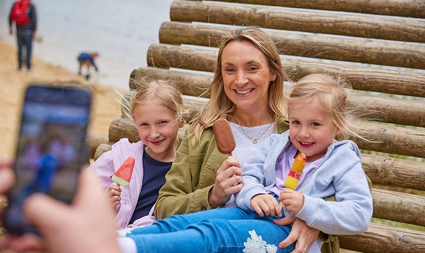 A woman with two children smile and hold ice creams while posing for a smartphone photo, sitting on a log bench at a beach, water and people in the background.