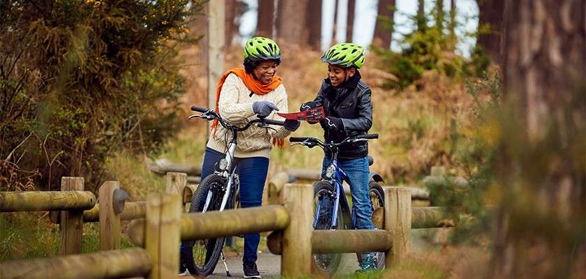 Two cyclists pause, smiling and examining a smartphone together while straddling bikes. They wear bright helmets and jackets. Wooden rails border a forest path with tall trees and shrubs.