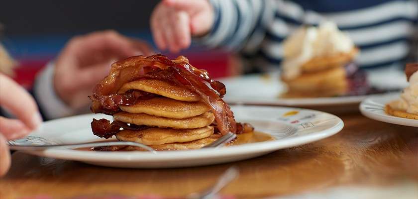 Pancake stack with crispy bacon, glossed with syrup, sits on a white plate as a child’s hand reaches; other plates of pancakes surround it on a wooden breakfast table.