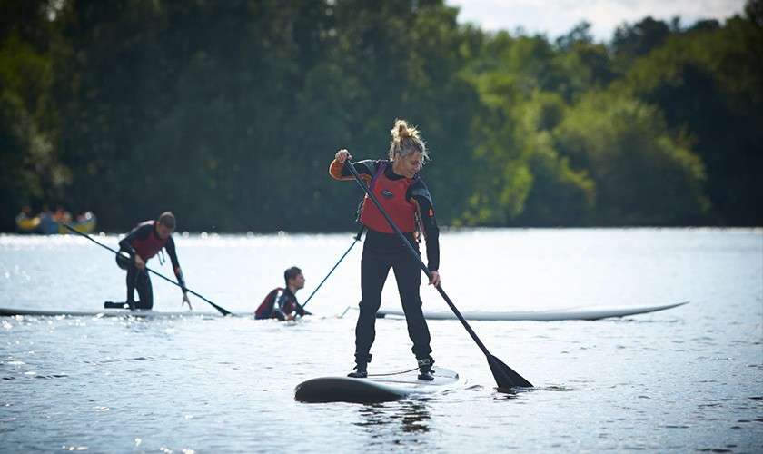 Person in wetsuit and red vest stands on a paddleboard, stroking forward with a long paddle; nearby paddlers kneel and balance on boards across a calm lake bordered by trees.