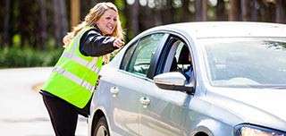 Traffic worker in a bright safety vest gestures and points, guiding a silver sedan with its window down along a road, with trees and sunlight in the background.
