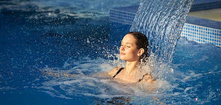Woman in a black swimsuit relaxes under a waterfall spout, eyes closed, as water cascades onto her head in a blue-tiled swimming pool.