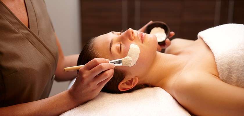Client’s face receives a creamy facial mask, applied with a brush by a practitioner; the client lies on a treatment bed under a white towel in a softly lit spa room.