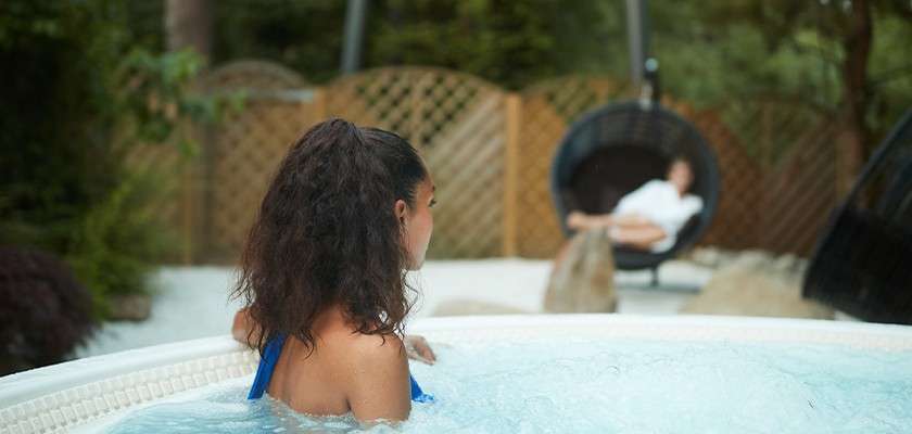 Woman in a blue swimsuit soaks in a bubbling hot tub, facing away. In the background, another person lounges in a hanging chair amid rocks, lattice fencing, and greenery.