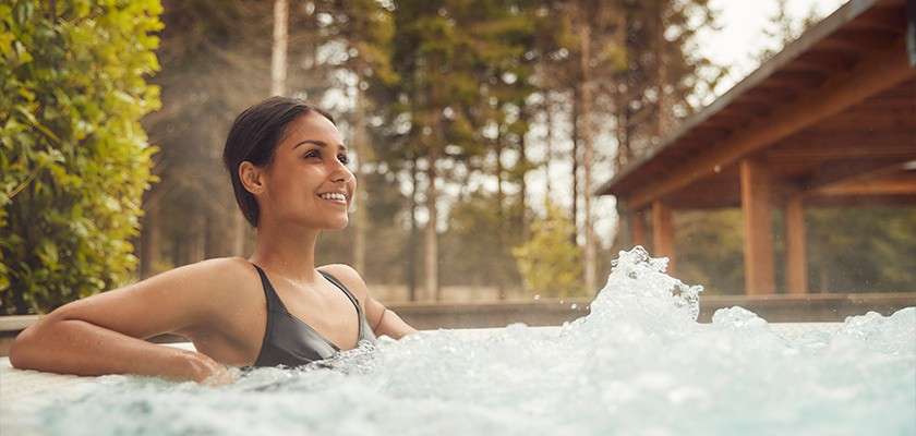 Woman relaxes, smiling in a bubbling outdoor hot tub, leaning on the edge; steam rises around her amid trees and a wooden pavilion in a serene, forested setting.