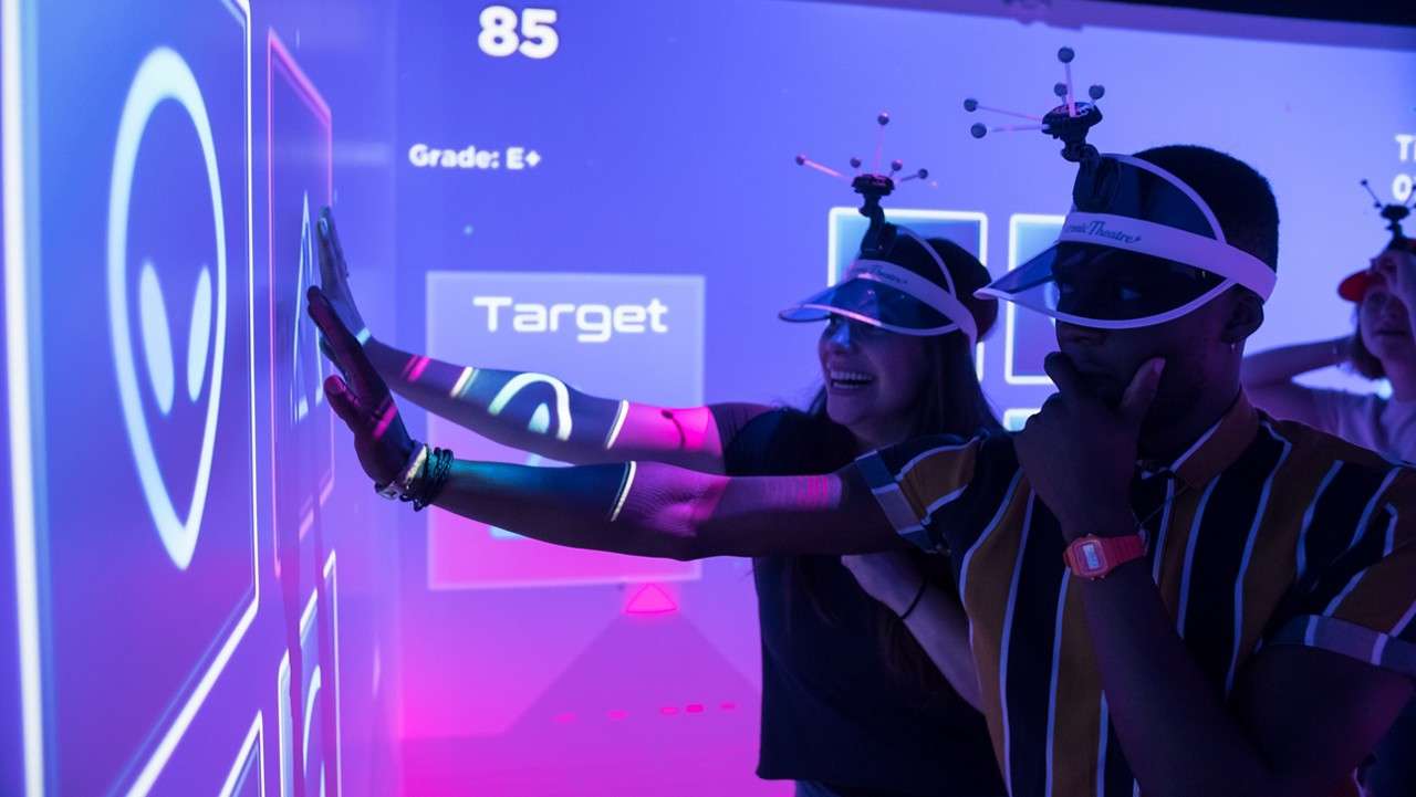 Players press glowing panels on a neon projection wall; they wear visors with tracking markers in a dim arcade. On-screen text: 85, Grade: E+, Target.