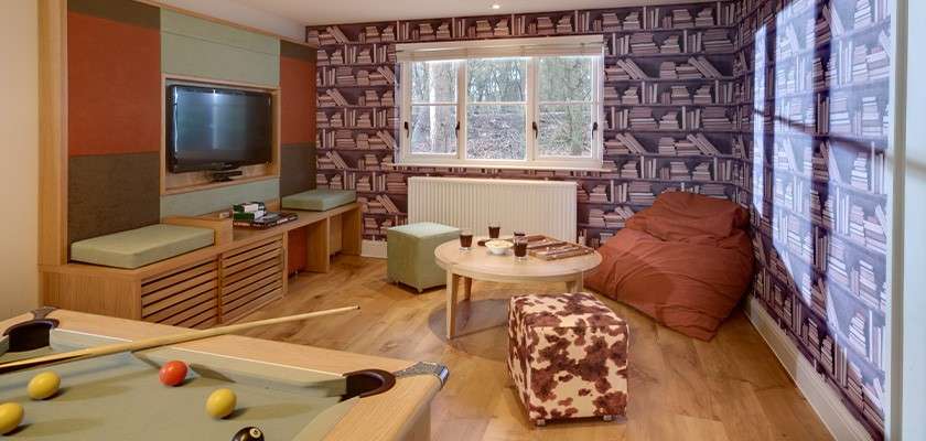 Pool table sits ready, balls scattered, in a cozy recreation room; television faces built-in benches, round coffee table with cups beside beanbag and ottoman; book-themed wallpaper and window overlook trees.