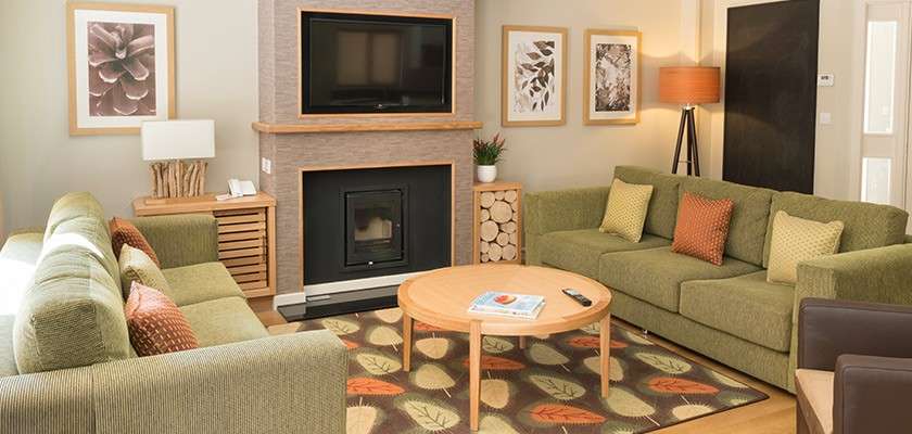 Green sofas encircle a round wooden coffee table, holding a remote and magazine, in a cozy living room with fireplace, wall-mounted TV, leaf-pattern rug, artwork, and warm lighting.