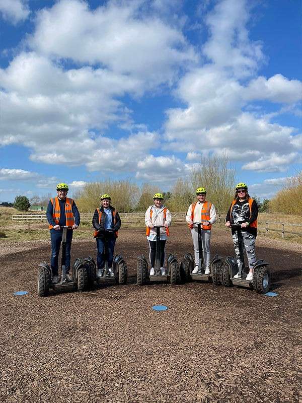 Five riders standing on Segways pose on a mulch track at an outdoor training area, wearing helmets and safety vests, with fences, sparse trees, and a blue, partly cloudy sky.