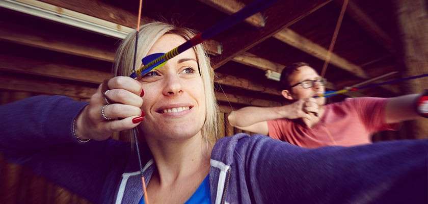 Two adults with bows draw arrows and aim forward, focusing downrange. They stand side by side in a rustic, roofed archery range with wooden beams, casual clothes, and concentrated expressions.
