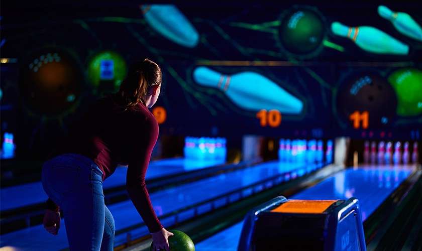 Bowler prepares to roll a green ball; pins glow ahead amid neon graphics in a dark bowling alley. Visible text: 10, 11.