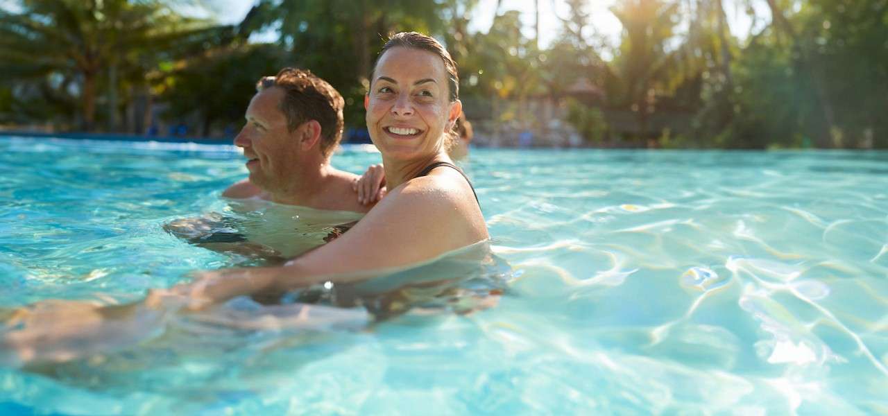 Two adults smile while wading chest-deep, one facing the camera, in a sunlit swimming pool; rippling turquoise water surrounds them, with palm trees and bright afternoon light in the background.