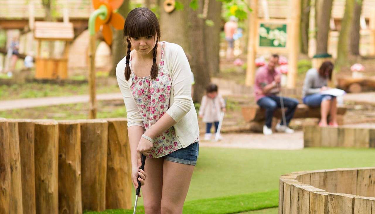 Young woman putts a mini-golf ball, concentrating, on an outdoor course with wooden barriers and artificial turf; a child and seated adults watch in a park-like setting.