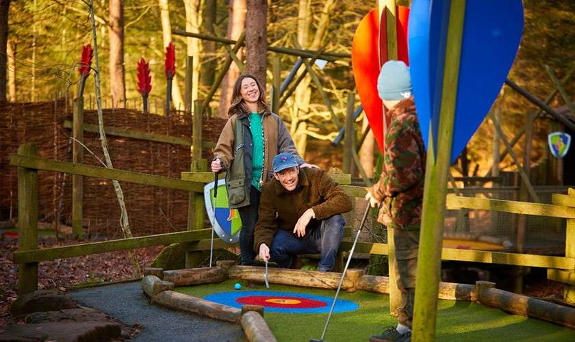 Three people play mini-golf; one crouches, putting a blue ball toward a target circle while others watch and smile; wooded outdoor course with wooden rails and colorful leaf-shaped obstacles.
