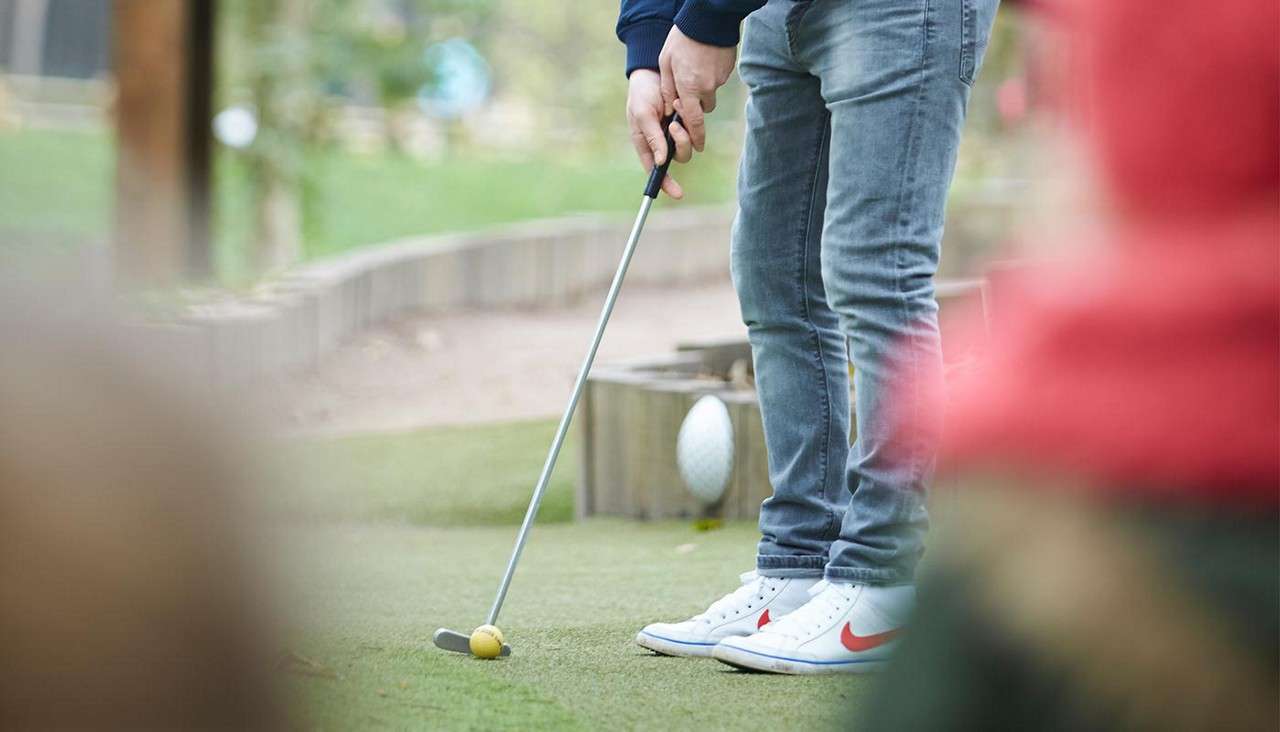 Golfer’s lower body aims a putter at a yellow ball, preparing to strike on an outdoor mini-golf course with artificial turf, wooden edging, and blurred spectators in the foreground.