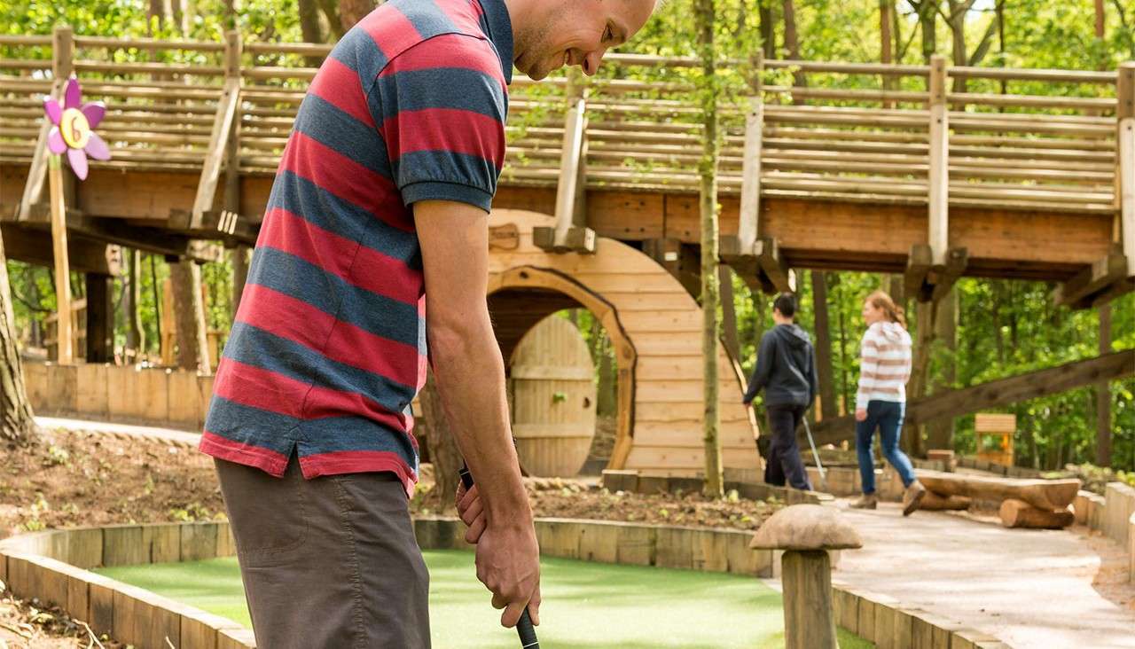 Golfer putts on a mini-golf green, aiming toward a wooden tunnel while others walk nearby; elevated wooden bridge and forest surround. Sign reads "6" on a flower-shaped marker.