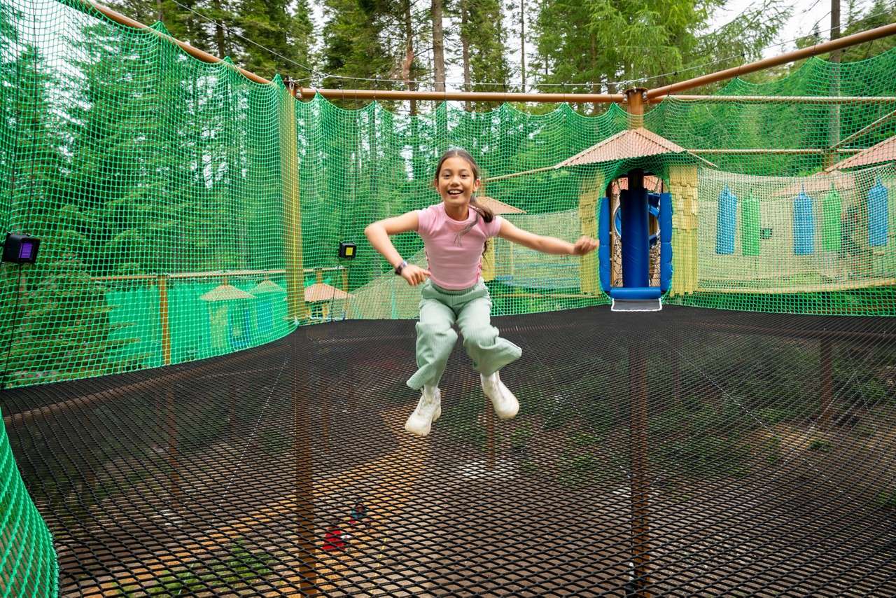 Child jumps midair on a suspended net trampoline, surrounded by green safety nets in a treetop adventure playground with hut-like platforms and a blue tube slide among tall trees.