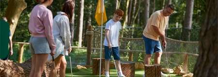 Golfer putts a ball as a boy watches; two others wait with clubs nearby, on a forested mini-golf course with rope fencing and wooden stump obstacles.