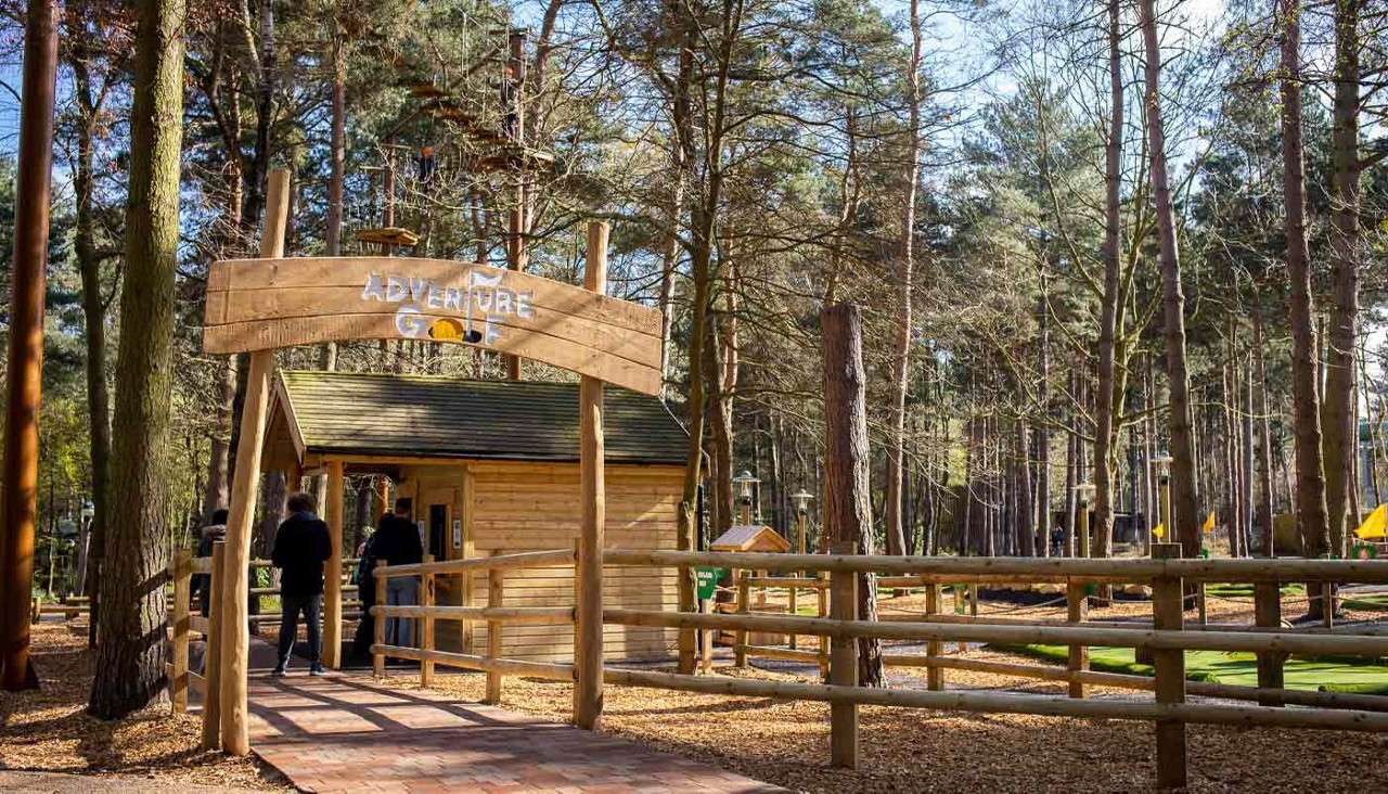 Wooden entrance sign reading ADVENTURE GOLF arches above a small hut, guiding visitors walking along a fenced path; set within a pine forest surrounding a mini-golf course.