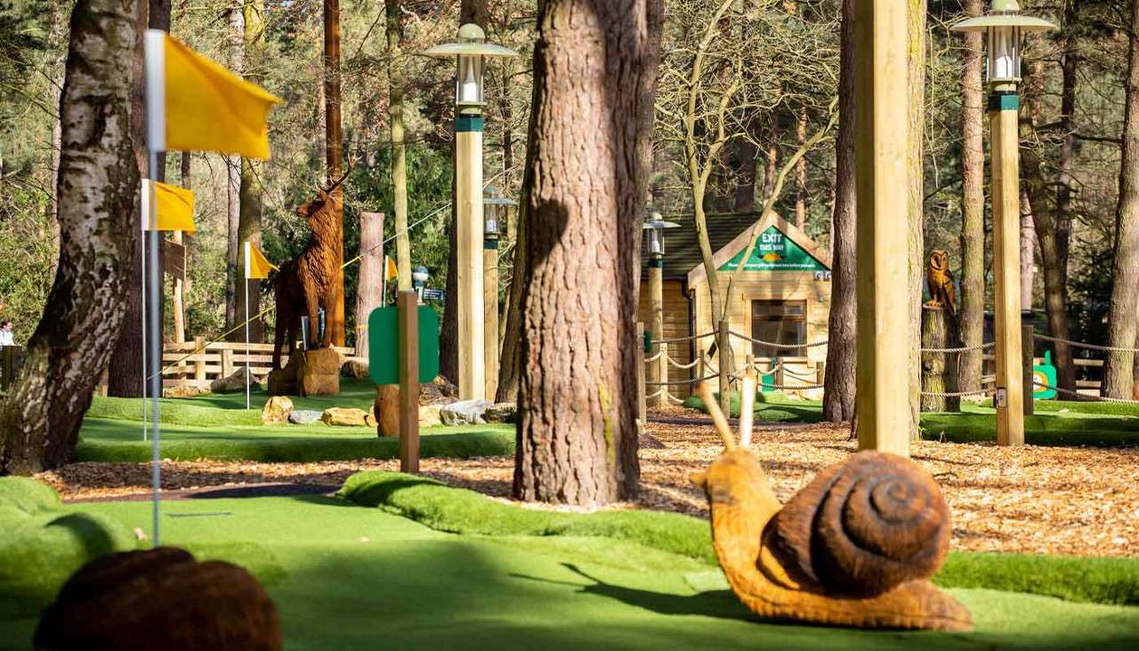 Wooden snail sculpture sits on artificial putting green, amid yellow-flagged mini-golf holes; surrounded by trees, lamp posts, and animal carvings near a small cabin signed: "EXIT THIS WAY".