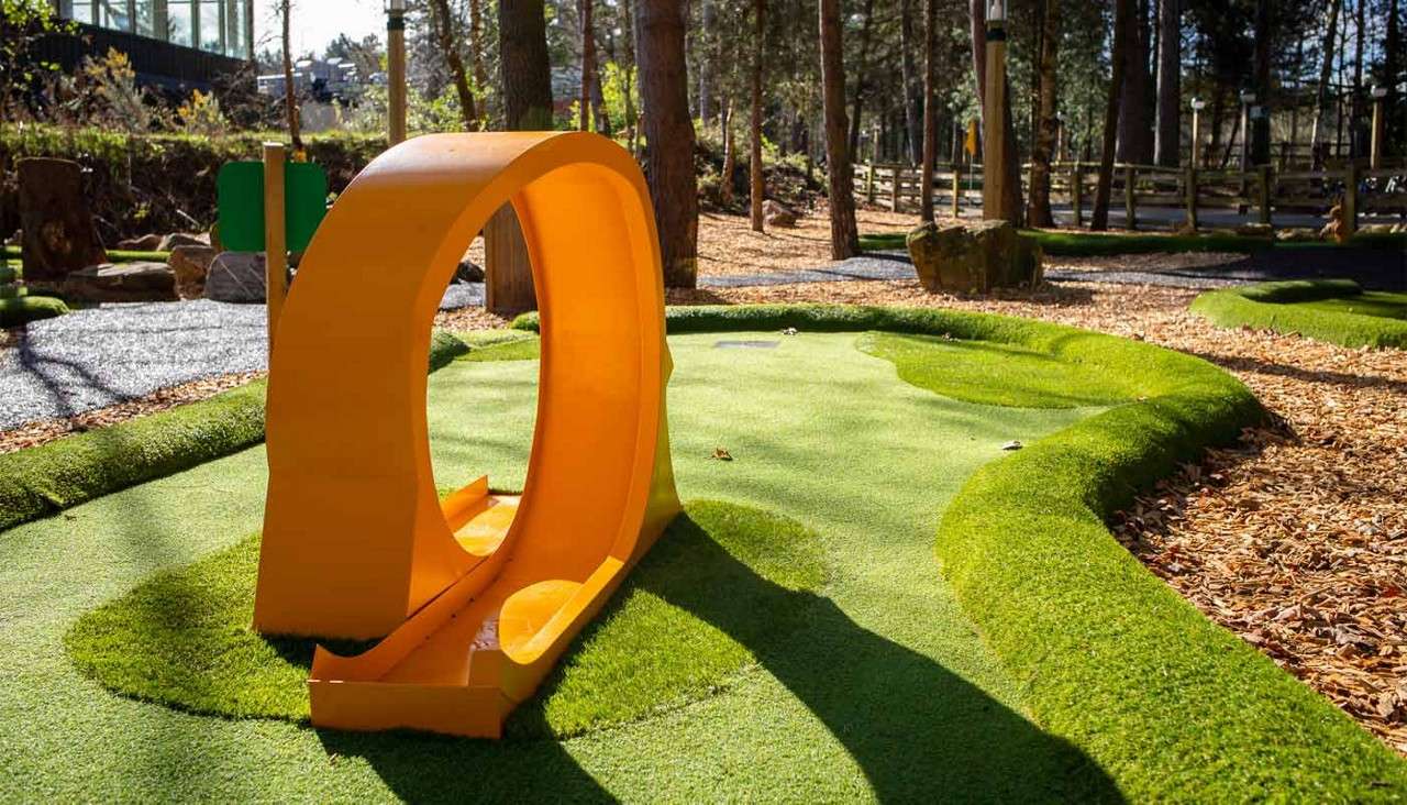 Orange loop obstacle directs a golf ball through a channel, positioned on undulating artificial turf within an outdoor mini-golf course surrounded by trees, wood-chip paths, rocks, and a fence.