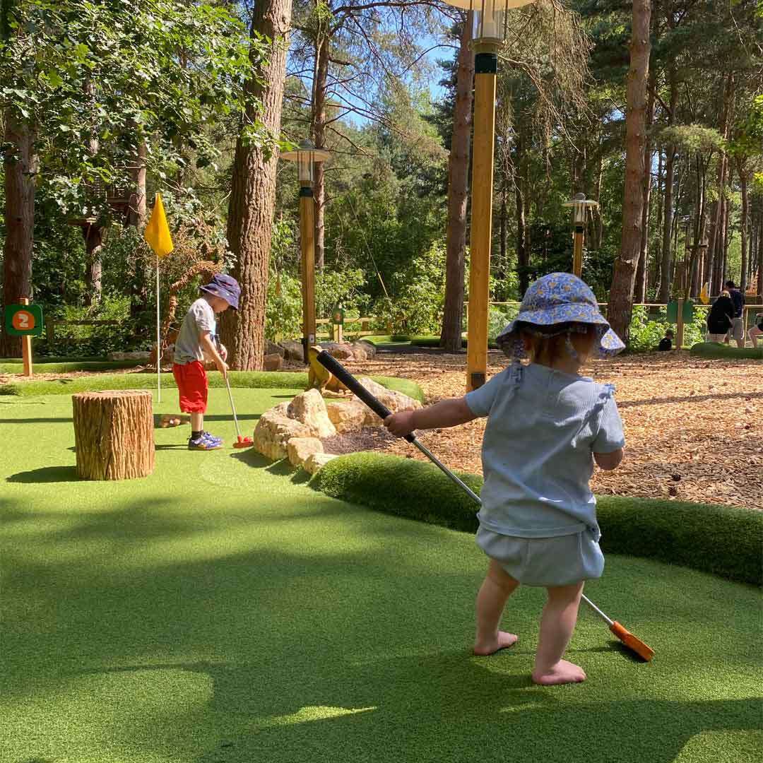 Two young children hold mini-golf putters; one putts, the other walks barefoot. Context: woodland mini-golf course with artificial turf, rocks, stump obstacle, hole flag, and a sign reading "2".