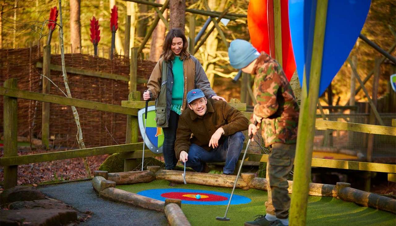 Child golfer putts a blue ball toward a colorful target hole, while two adults watch and guide, in an outdoor mini-golf course surrounded by wooden fences, trees, and playful decorations.