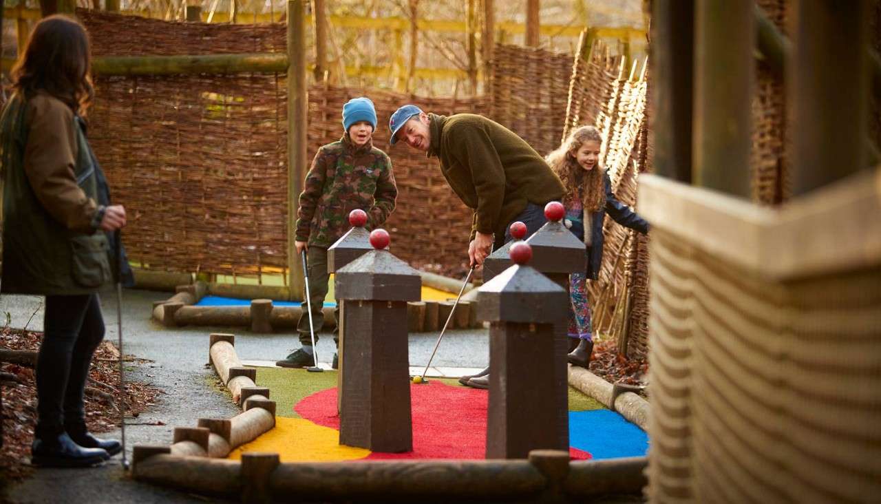 Man putts a mini-golf ball while two children and another adult watch, smiling. The game unfolds on an outdoor course with colorful turf and wooden obstacles, enclosed by woven fences.