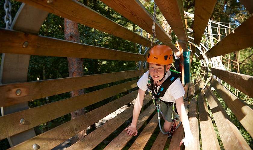 Climber crawls through a wooden slat tunnel, clipped to an overhead safety line, wearing an orange helmet labeled "Petzl." Trees and a ropes course surround them, with another participant following.