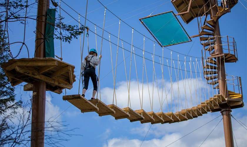 A harnessed person crosses a suspended rope bridge of wooden planks, gripping safety lines, within a high-ropes course featuring platforms, cables, and a spiral staircase against a blue, cloudy sky.